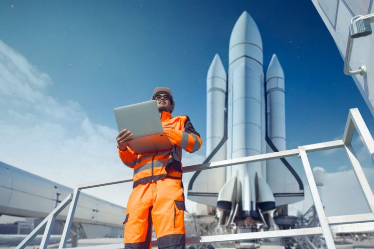 Engineer in orange high vis clothing holding laptop stood in front of a space rocket.