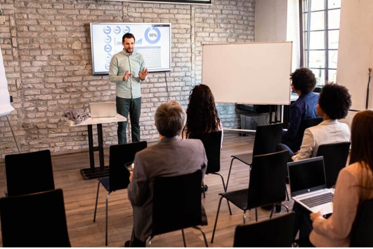 Person stood in front of a whiteboard looking towards a group of people sat in chairs looking forward.