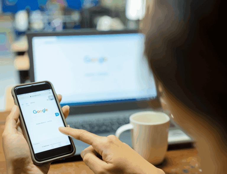 Person sat at computer with Google search engine displayed on monitor and mobile phone.